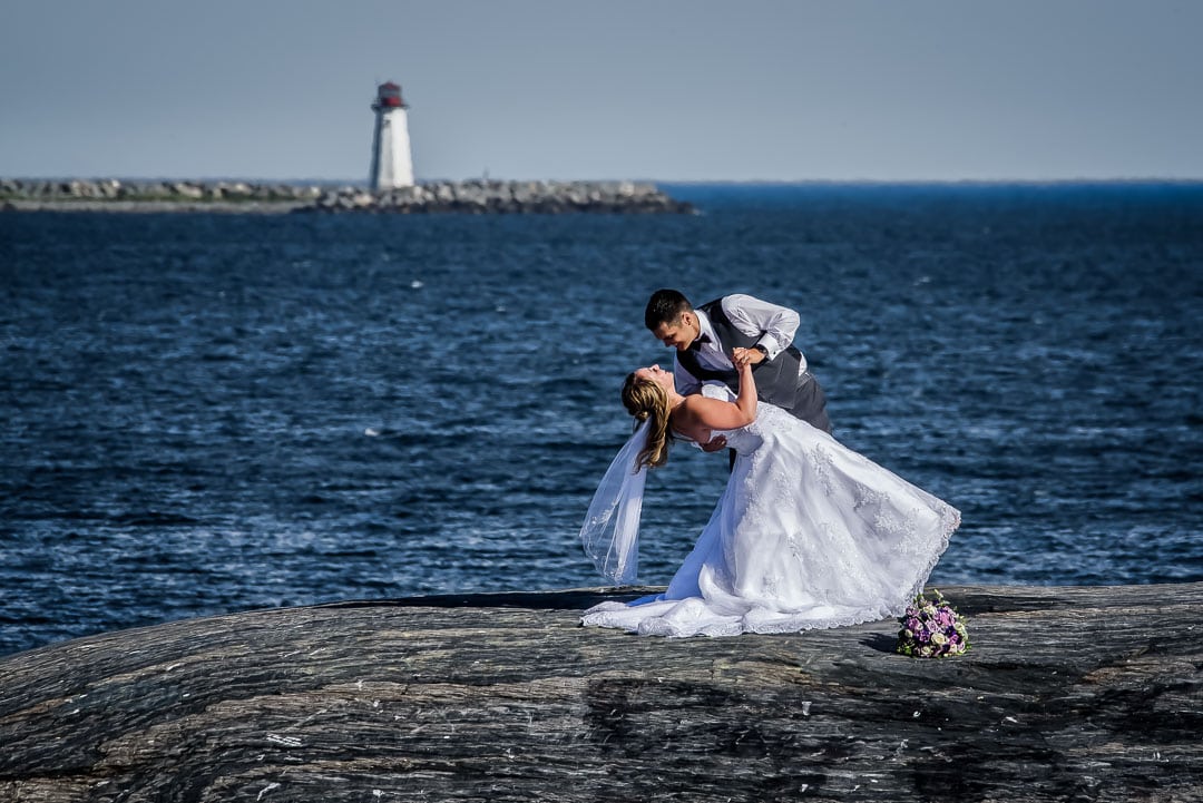The groom dips the bride for wedding photos in front of a lighthouse at the Point Pleasant Park, captured by Halifax wedding photographers.