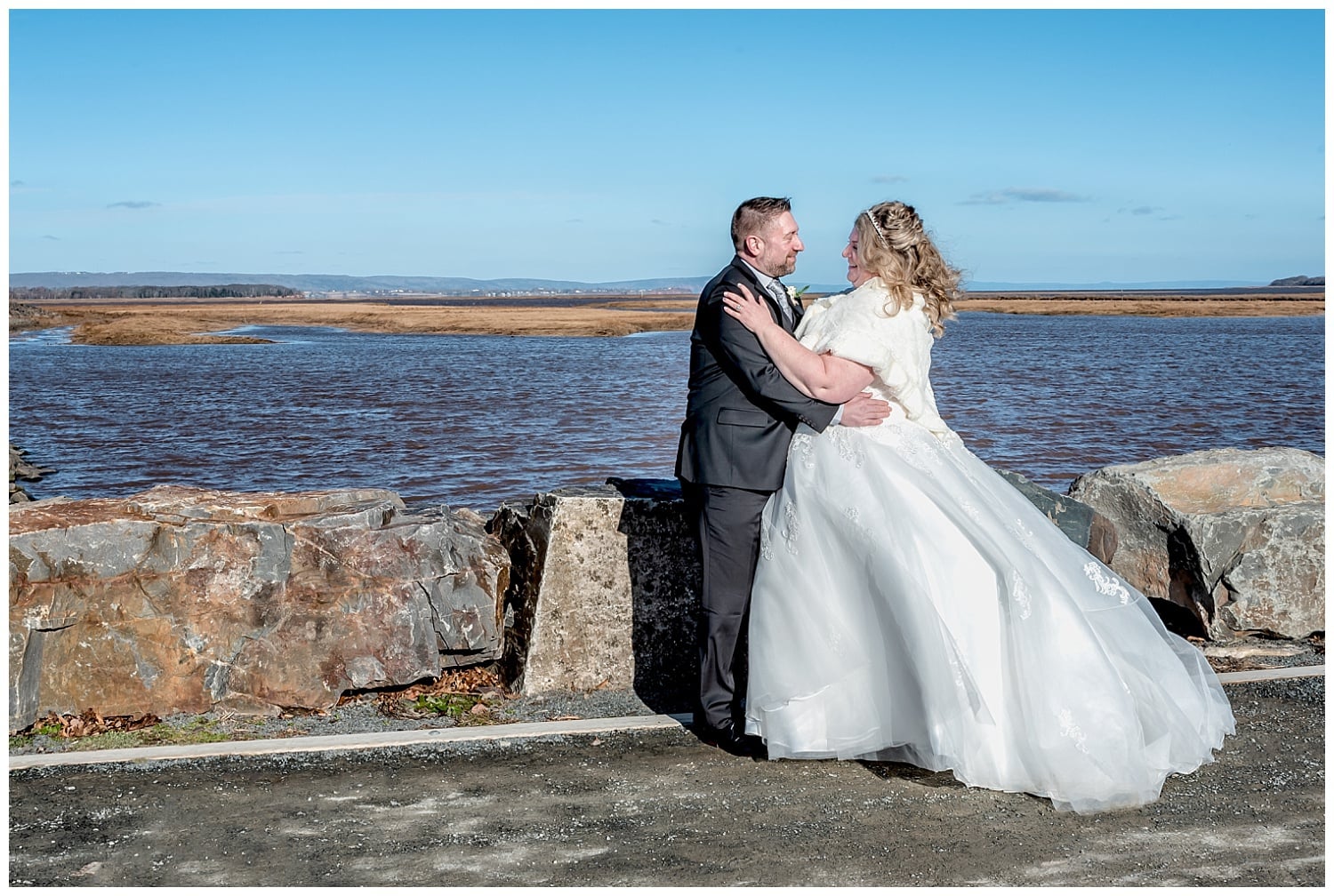 The bride and groom pose for wedding photos on the ocean behind the Wolfville Chapel in Nova Scotia.