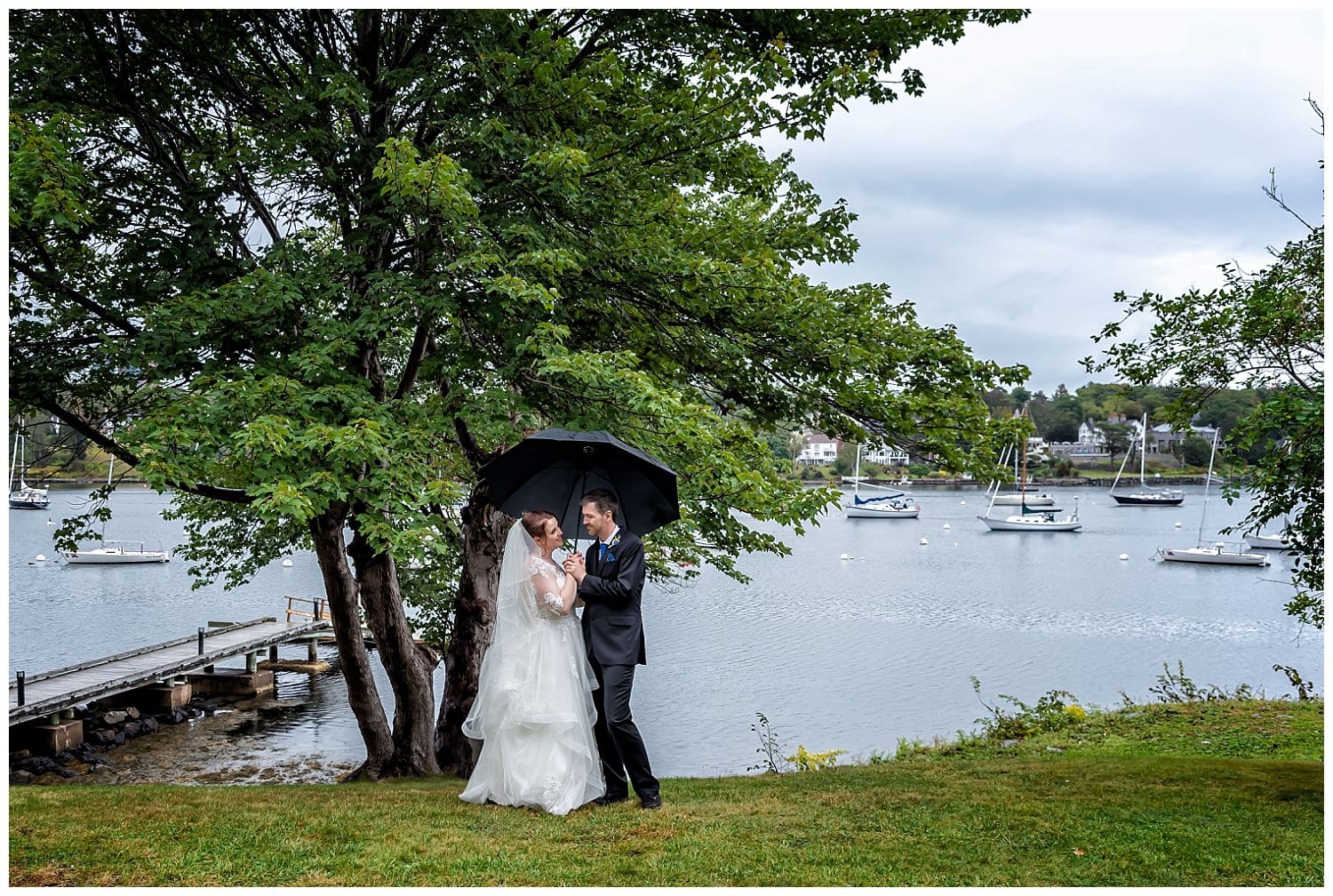 The bride and groom stand along the waterfront at Saraguay House for wedding photos under an umbrella.