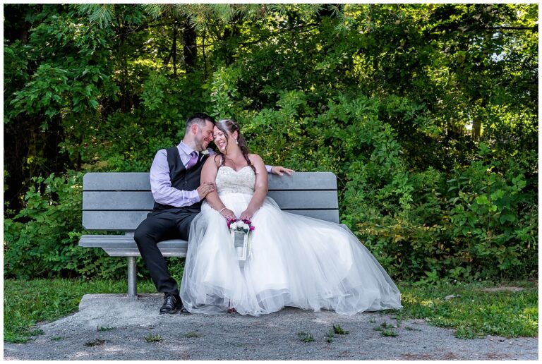 The bride and groom sit on a bench for their wedding photos at First Park in Sackville NS.