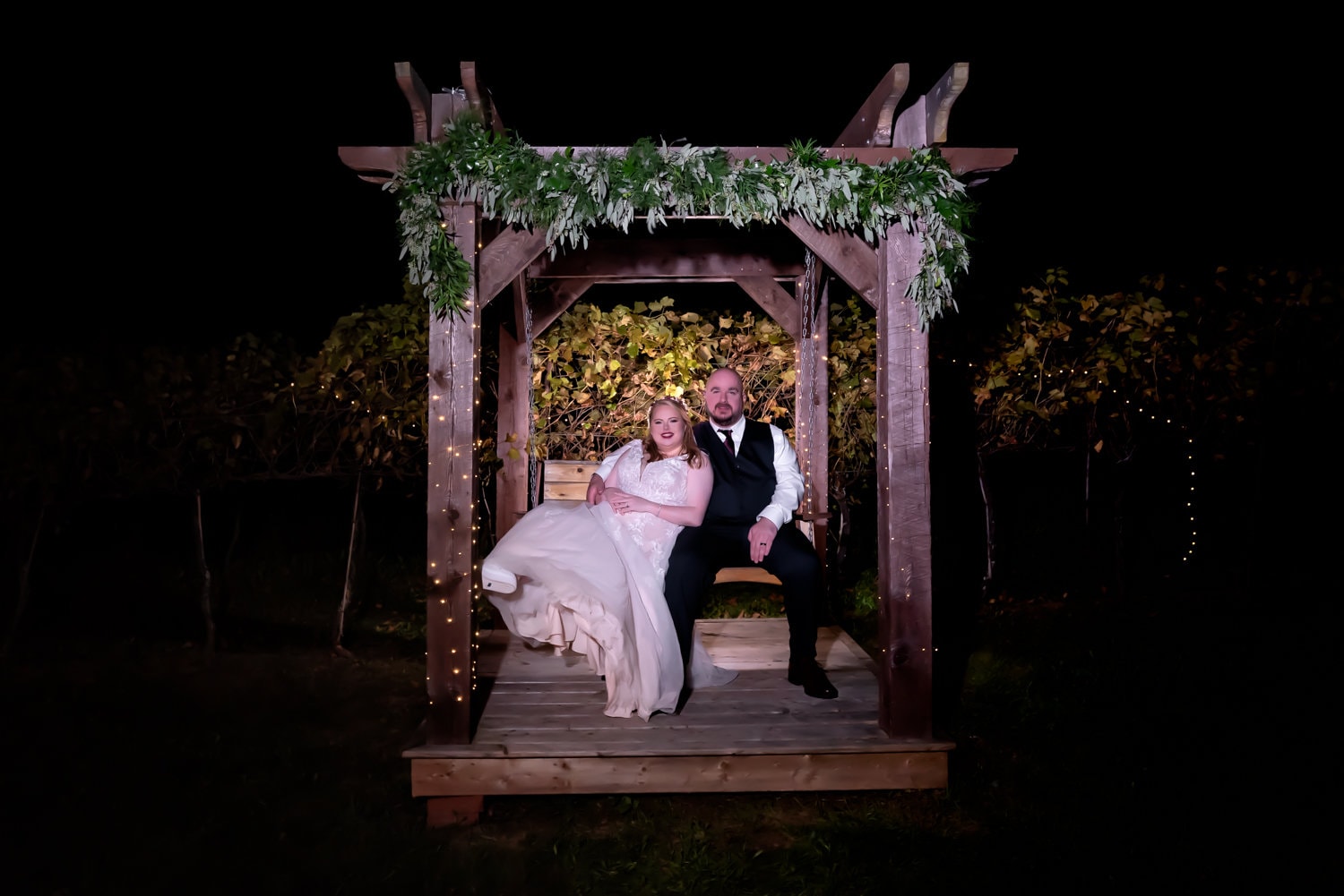 The bride and groom sit on a wooden swing and pose f or