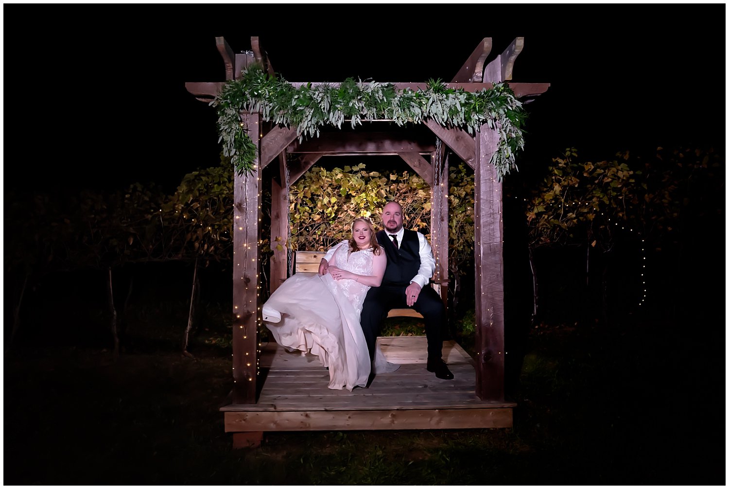 The bride and groom pose on a swing at night infront of mini lights at the Bent Ridge Winery in NS.