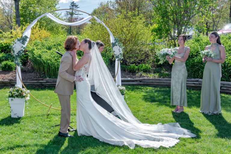 Bride in a white gown and groom in a tan suit standing under a floral arch having their first kiss during their outdoor ceremony at the Ashburn Golf Club in Halifax.
