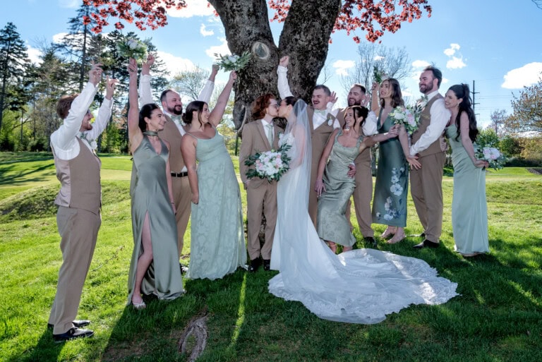Wedding party gathered under a red‑leafed tree as the bride and groom share a kiss.