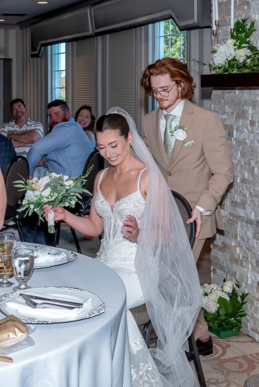 Bride getting seated with her bouquet while the groom stands behind her to push the chair in during their Ashburn Golf Club reception.