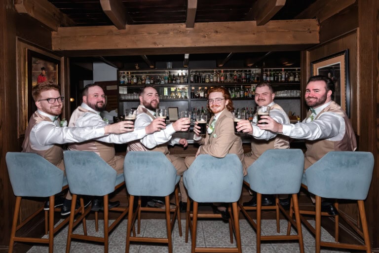 Groomsmen sitting at a rustic bar, smiling and raising glasses of dark beer before the wedding at the Lord Nelson Hotel in Halifax Nova Scotia.