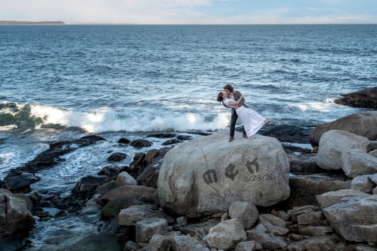 Kage dips Megan for their engagement photos by the ocean, standing on a massive rock with their wedding date etched into it.