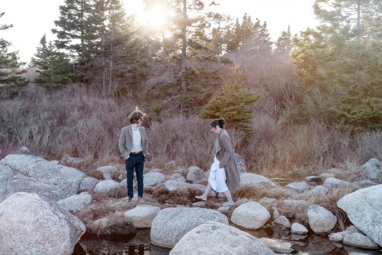 Engagement photo of a couple exploring a wooded area with ocean rocks in Halifax Nova Scotia with warm golden light filtering through the trees.