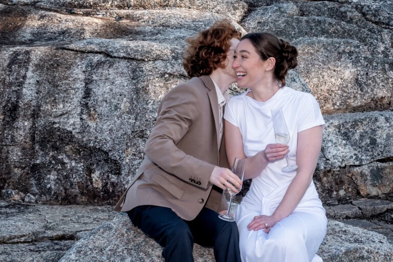 Couple sitting on rocky terrain by the ocean sharing a champagne toast during their engagement session in Halifax Nova Scotia.