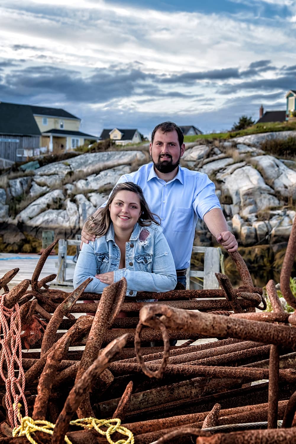 Engagement Portfolio Page A newly engaged couple pose for engagement photos infront of rusty anchors at Peggys Cove Nova Scotia.