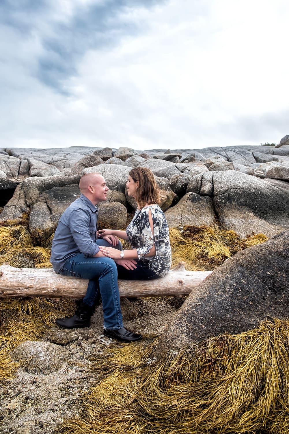 Engagement Portfolio Page A love couple face each other holding hands sitting on a log at Peggys Cove for their engagement photos.